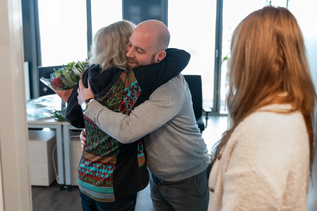 Two colleagues sharing a warm hug in the office, one person holding a bouquet of flowers